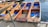 Wooden boats with blue cushions are aligned on a dock, chained and ready for use on the water. Each boat has a unique identification number and a paddle rests across one of them.