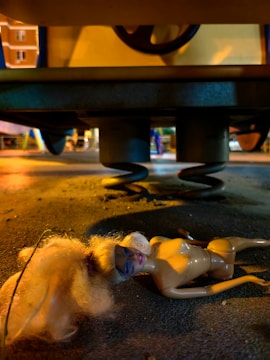 A doll is lying on the ground beneath a large structure with a metal spring. The scene is illuminated by artificial yellow light, creating a dramatic shadow and highlighting the doll's worn appearance and disheveled hair. In the background, buildings and blurred objects suggest an urban environment.
