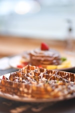 Close-up of waffles drizzled with chocolate sauce on a plate, with a side of pancakes topped with a strawberry in the background. The setting appears to be a breakfast or brunch table with soft lighting.