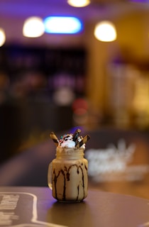 A jar filled with a creamy beverage, topped with whipped cream, colorful sprinkles, and chocolate sticks. The jar has chocolate syrup drizzled down its sides. It is placed on a blurred background of a warmly lit interior with soft colorful lights.