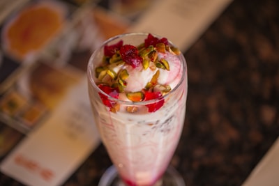 A tall glass filled with a creamy dessert, topped with pistachios and a red syrup. The glass contains layers of ingredients, likely including ice cream or yogurt, with red and white swirls. The setting is casual with blurred background elements, suggesting a dining table.