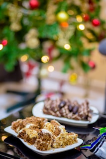 Two plates of assorted pastries are placed on a reflective table surface. The background features a beautifully decorated Christmas tree with colorful ornaments and blurred festive lights.