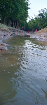 A shallow stream with gentle water flow runs through a natural setting bordered by trees. The ground alongside the stream is earthy and slightly muddy. In the background, several people are visible, giving a sense of human presence within the landscape.