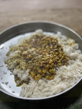 A metal plate filled with white rice topped with cooked lentils displaying a mixture of yellows and browns. The lentils appear to be seasoned and have a glossy texture, indicating possible oil or sauce.