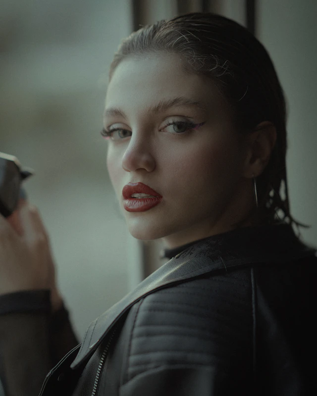 Margaux by a window in a leather jacket, phone in hand, glancing over her shoulder.