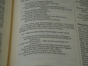 A close-up view of an open book page from the Bible, showing printed text in English. The text is clearly legible and features biblical verses with some words in bold and small numbers indicating verse numbers.