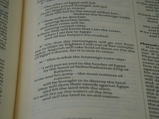 A close-up view of an open book page from the Bible, showing printed text in English. The text is clearly legible and features biblical verses with some words in bold and small numbers indicating verse numbers.