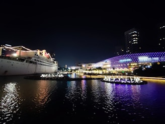 A large cruise ship is illuminated with strings of lights, docked near a waterfront cityscape at night. The water reflects the ship and city lights. The background features a modern building with vibrant purple and blue light patterns, showcasing a lively and colorful atmosphere.