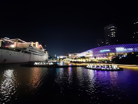 A large cruise ship is illuminated with strings of lights, docked near a waterfront cityscape at night. The water reflects the ship and city lights. The background features a modern building with vibrant purple and blue light patterns, showcasing a lively and colorful atmosphere.