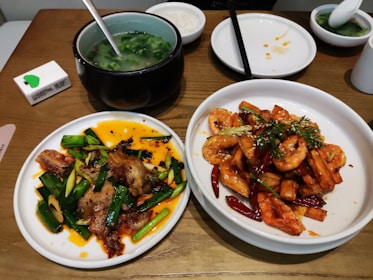 A wooden table set with several dishes, including a plate of fried pork and green onions in a spicy orange sauce, another plate with shrimp, dried red chilies, and herbs. There is also a bowl of green leafy soup, a small bowl with sauce, and a box of tissues.