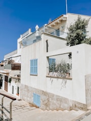 A white residential building with blue shutters and a rooftop terrace, set against a clear blue sky. The architecture features clean lines and minimalistic design. A small potted plant is visible on a windowsill, and a pathway runs alongside the building.