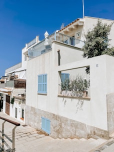 A white residential building with blue shutters and a rooftop terrace, set against a clear blue sky. The architecture features clean lines and minimalistic design. A small potted plant is visible on a windowsill, and a pathway runs alongside the building.