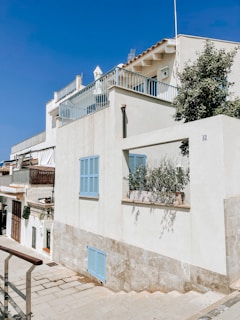 A white residential building with blue shutters and a rooftop terrace, set against a clear blue sky. The architecture features clean lines and minimalistic design. A small potted plant is visible on a windowsill, and a pathway runs alongside the building.