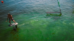 A person is balancing on a hydrofoil board gliding over calm green water. A buoy is visible in the distance, and there is a partially submerged ramp nearby. The sunlight creates reflections and patterns on the water surface.