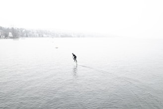 A person is hydrofoiling across a vast body of water surrounded by a foggy environment. The water is calm, and the person appears to be in a wetsuit, gliding smoothly. The background is shrouded in mist, obscuring the distant shoreline and trees.