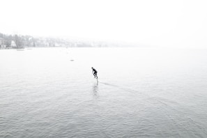 A person is hydrofoiling across a vast body of water surrounded by a foggy environment. The water is calm, and the person appears to be in a wetsuit, gliding smoothly. The background is shrouded in mist, obscuring the distant shoreline and trees.