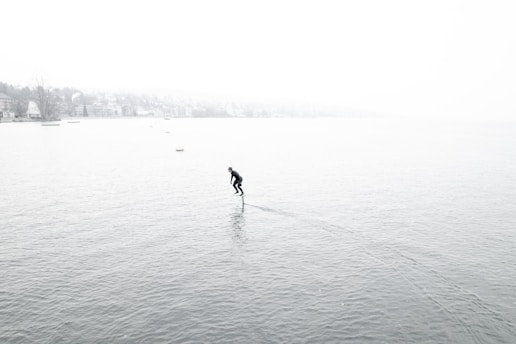 A person is hydrofoiling across a vast body of water surrounded by a foggy environment. The water is calm, and the person appears to be in a wetsuit, gliding smoothly. The background is shrouded in mist, obscuring the distant shoreline and trees.