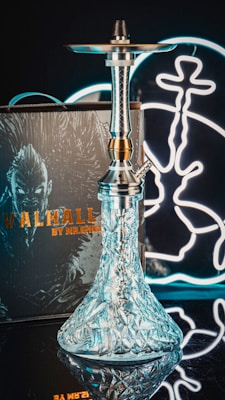 A modern hookah with a textured glass base and metallic top stands in the foreground. The backdrop includes a neon light with abstract designs and a box labeled 'Valhalla by Mr. Shisha' featuring an intricate illustration. The scene combines elements of traditional and contemporary aesthetics.