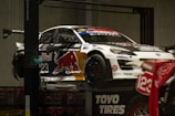 A race car adorned with various sponsor logos, including Red Bull, is elevated on a platform. The car is predominantly white with contrasting dark elements and a large red bull graphic on the side. Nearby, there is a motorcycle with the number 123 visible. The setting appears to be indoors, with concrete walls and a subdued lighting ambiance.