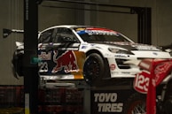 A race car adorned with various sponsor logos, including Red Bull, is elevated on a platform. The car is predominantly white with contrasting dark elements and a large red bull graphic on the side. Nearby, there is a motorcycle with the number 123 visible. The setting appears to be indoors, with concrete walls and a subdued lighting ambiance.