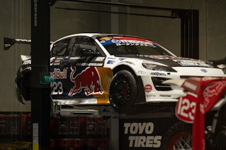 A race car adorned with various sponsor logos, including Red Bull, is elevated on a platform. The car is predominantly white with contrasting dark elements and a large red bull graphic on the side. Nearby, there is a motorcycle with the number 123 visible. The setting appears to be indoors, with concrete walls and a subdued lighting ambiance.