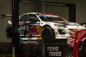 A race car adorned with various sponsor logos, including Red Bull, is elevated on a platform. The car is predominantly white with contrasting dark elements and a large red bull graphic on the side. Nearby, there is a motorcycle with the number 123 visible. The setting appears to be indoors, with concrete walls and a subdued lighting ambiance.