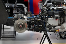 A close-up of a complex mechanical assembly, showcasing a car's braking and engine system, including visible components such as a brake disc with red Brembo branding, various hoses, pipes, and parts of a motor. The setting appears to be an automotive workshop or garage with other equipment in the background.