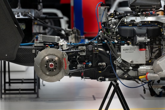 A close-up of a complex mechanical assembly, showcasing a car's braking and engine system, including visible components such as a brake disc with red Brembo branding, various hoses, pipes, and parts of a motor. The setting appears to be an automotive workshop or garage with other equipment in the background.
