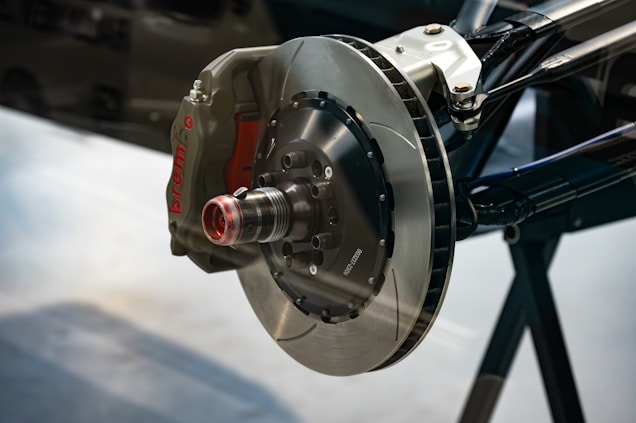 A close-up view of a brake disc and caliper system, highlighted by a shiny metallic rotor and a red caliper labeled with 'brembo'. The setup is mounted on a mechanical arm or part, featuring various bolts and components designed for high-performance applications. The background is blurred, focusing attention on the intricate details and design of the braking system.