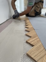 A person wearing gloves is installing wooden flooring. The focus is on the zigzag pattern of the wood pieces being laid down onto a layer of adhesive.