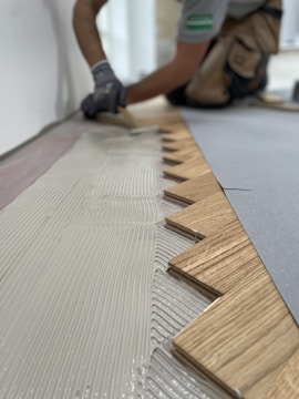 A person wearing gloves is installing wooden flooring. The focus is on the zigzag pattern of the wood pieces being laid down onto a layer of adhesive.