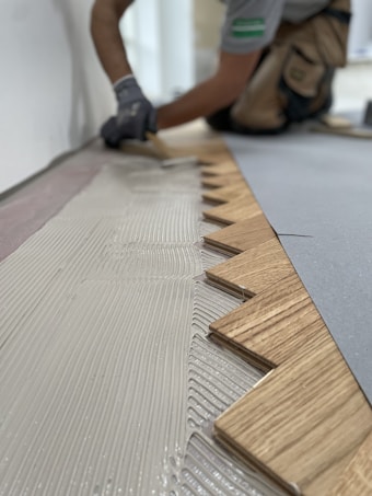 A person wearing gloves is installing wooden flooring. The focus is on the zigzag pattern of the wood pieces being laid down onto a layer of adhesive.