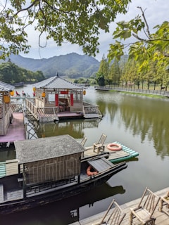 A serene lake scene featuring two traditional boats with covered seating and decorative elements, floating gently on calm water. Surrounding the lake are lush green trees and a mountainous backdrop. A few chairs are placed on a wooden deck, with life buoys nearby for safety. The sky is slightly cloudy, offering a peaceful and tranquil atmosphere.