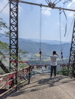 A person is standing on a scenic lookout platform, taking a photograph or observing the breathtaking view of a vast landscape filled with mountains and greenery. The platform is decorated with hanging red ornaments, possibly charms or wishes. The bright sky and distant mountains create a serene backdrop.
