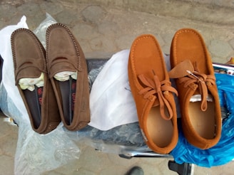 Two pairs of shoes are displayed on a motorcycle seat, one brown pair of loafers and one orange pair of lace-up shoes. They are placed on plastic bags, with the pavement visible in the background.