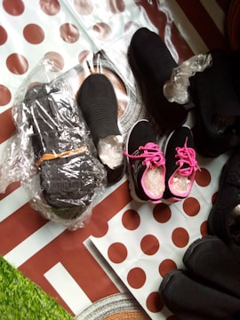 Several pairs of black shoes are arranged on a surface covered with polka dot paper. Some shoes are wrapped in plastic, and one pair features bright pink laces, creating a striking contrast. The setup includes hints of a green grassy texture.