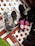 Several pairs of black shoes are arranged on a surface covered with polka dot paper. Some shoes are wrapped in plastic, and one pair features bright pink laces, creating a striking contrast. The setup includes hints of a green grassy texture.
