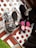 Several pairs of black shoes are arranged on a surface covered with polka dot paper. Some shoes are wrapped in plastic, and one pair features bright pink laces, creating a striking contrast. The setup includes hints of a green grassy texture.