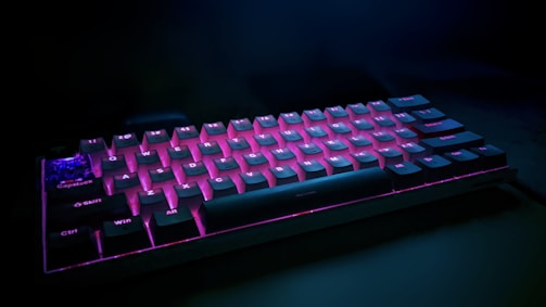 A mechanical keyboard with backlit keys displaying a vibrant purple and blue glow sits at an angle against a dark background. The keys have a sleek design with visible legends, and the lighting highlights the keyboard’s contoured structure.