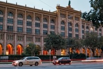 A neoclassical building with tall arched windows stands in the background, framed by trees. Warm orange lights illuminate the arches of the building, creating a striking contrast with the stone facade. In the foreground, a light-colored taxi with 'mytaxi' branding and a dark-colored car move along the street, capturing a sense of urban movement.