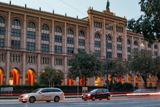 A neoclassical building with tall arched windows stands in the background, framed by trees. Warm orange lights illuminate the arches of the building, creating a striking contrast with the stone facade. In the foreground, a light-colored taxi with 'mytaxi' branding and a dark-colored car move along the street, capturing a sense of urban movement.