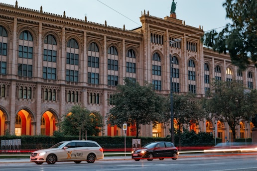 A neoclassical building with tall arched windows stands in the background, framed by trees. Warm orange lights illuminate the arches of the building, creating a striking contrast with the stone facade. In the foreground, a light-colored taxi with 'mytaxi' branding and a dark-colored car move along the street, capturing a sense of urban movement.