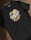 A black t-shirt with an illustrated design of a small space character lying on its side, wearing a helmet, positioned in the center. Surrounding the character are the words 'BE HAPPY' in bold, distressed yellow letters, with small circular accents.