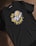 A black t-shirt with an illustrated design of a small space character lying on its side, wearing a helmet, positioned in the center. Surrounding the character are the words 'BE HAPPY' in bold, distressed yellow letters, with small circular accents.