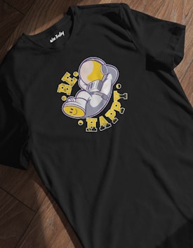 A black t-shirt with an illustrated design of a small space character lying on its side, wearing a helmet, positioned in the center. Surrounding the character are the words 'BE HAPPY' in bold, distressed yellow letters, with small circular accents.