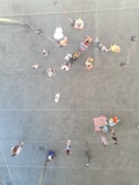 A group of people are reflected on a mirrored surface, creating an illusion of being above them. They walk on a paved area, some are visible with strollers, bags, and colorful clothes. Red and white barriers delineate a path on the ground.