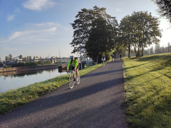 A group of cyclists ride along a paved path next to a calm river. The scene is set in a park-like environment with lush green grass and tall trees lining the pathway. The early morning sun casts long shadows and creates a peaceful and serene atmosphere.