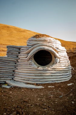 A structure composed of stacked, coiled sandbags forms a dome-like shape with a circular opening in the center. The background features a dry, hilly landscape under a clear blue sky. The ground is covered in dirt and small rocks, contributing to a rustic and rugged appearance.