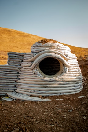 A structure composed of stacked, coiled sandbags forms a dome-like shape with a circular opening in the center. The background features a dry, hilly landscape under a clear blue sky. The ground is covered in dirt and small rocks, contributing to a rustic and rugged appearance.