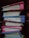 A stack of six thick notebooks or journals with colorful covers is arranged on a dark shelf. The spines are mostly visible, showing some signs of wear. The covers are of different colors including red, blue, and pink.